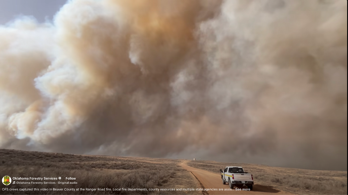 The image shows a vast landscape dominated by a massive cloud of smoke covering the sky. The smoke appears to be rising from a fire, creating billows of varying shades of gray and brown. In the foreground, there is a dirt road leading off into the distance, lined with dry grass on both sides. A white pickup truck is parked on the road, with its rear facing the viewer. The overall scene conveys a sense of impending danger due to the approaching smoke.