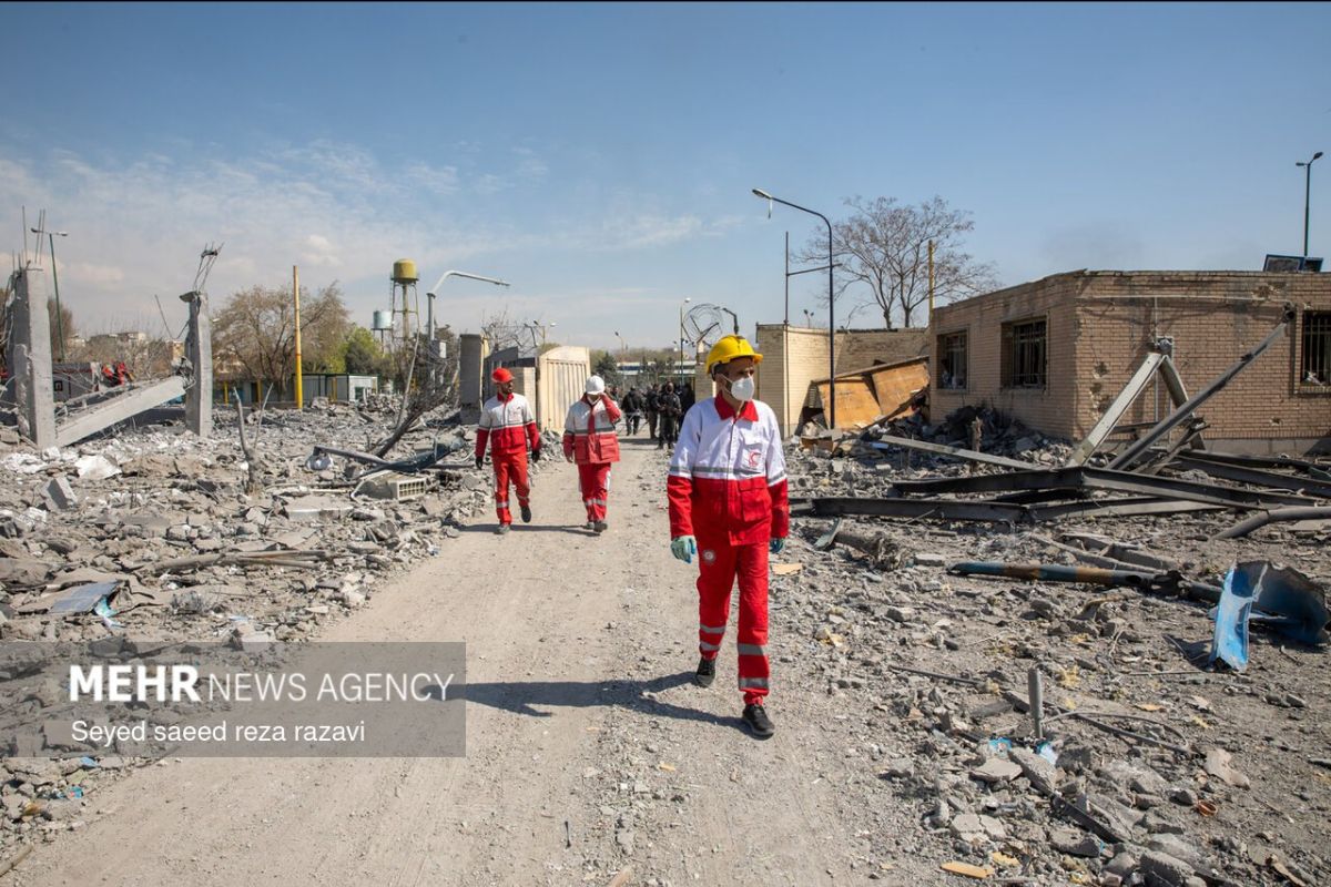 The image depicts a scene of devastation with debris and rubble spread across the ground, indicating the aftermath of a destructive event. Three individuals dressed in red and white uniforms walk along a dirt path amidst the wreckage. One of them is wearing a yellow helmet and a mask. Surrounding the path are damaged buildings, including a partially collapsed structure on the left and a brick building on the right. In the background, there are bare trees, a water tower, and several street lamps. The sky is clear with minimal clouds.