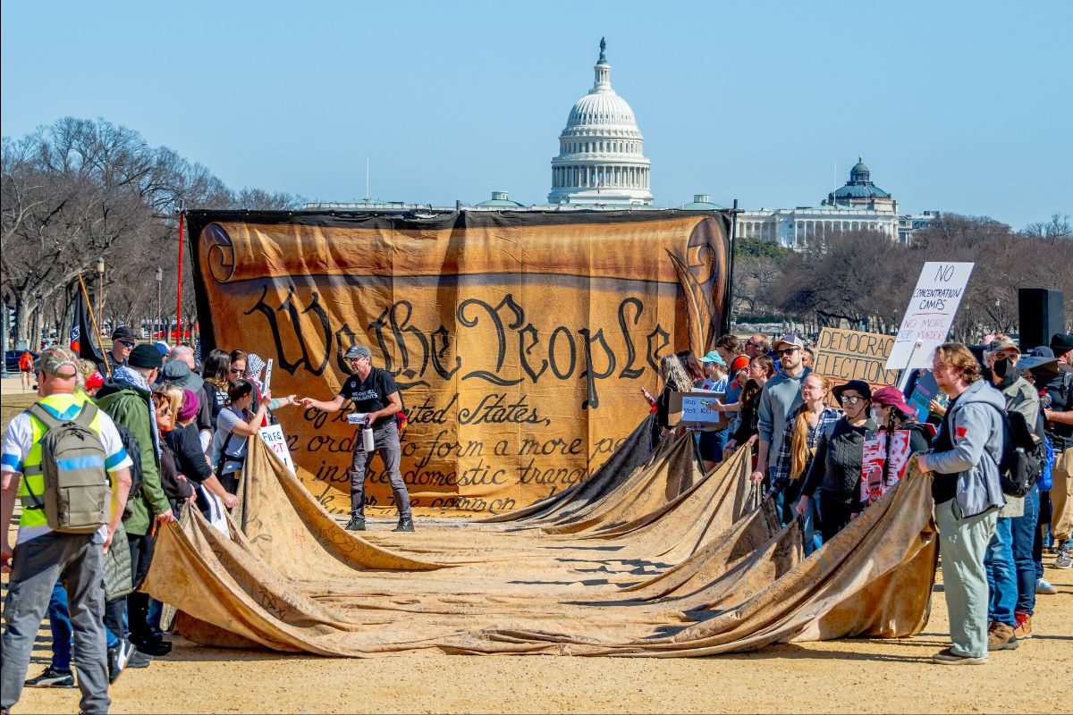 The image depicts a large gathering of people outdoors on a sunny day, in front of a giant replica of the United States Constitution's preamble with the words "We the People" prominently displayed. The replica covers a significant portion of the scene and is held up by several participants. In the background, the U.S. Capitol building is visible with its distinctive white dome, accompanied by other government buildings. The crowd, composed of individuals in casual attire, holds various signs. Some signs include messages such as "NO MORE STOLEN ELECTIONS" and "DEMOCRACY NOT AUTOCRACY." The setting appears to be a political demonstration or protest on a large expanse of sandy, open ground, typical of an area like the National Mall in Washington, D.C. The season is likely late fall or early spring, as the surrounding trees are bare without leaves.