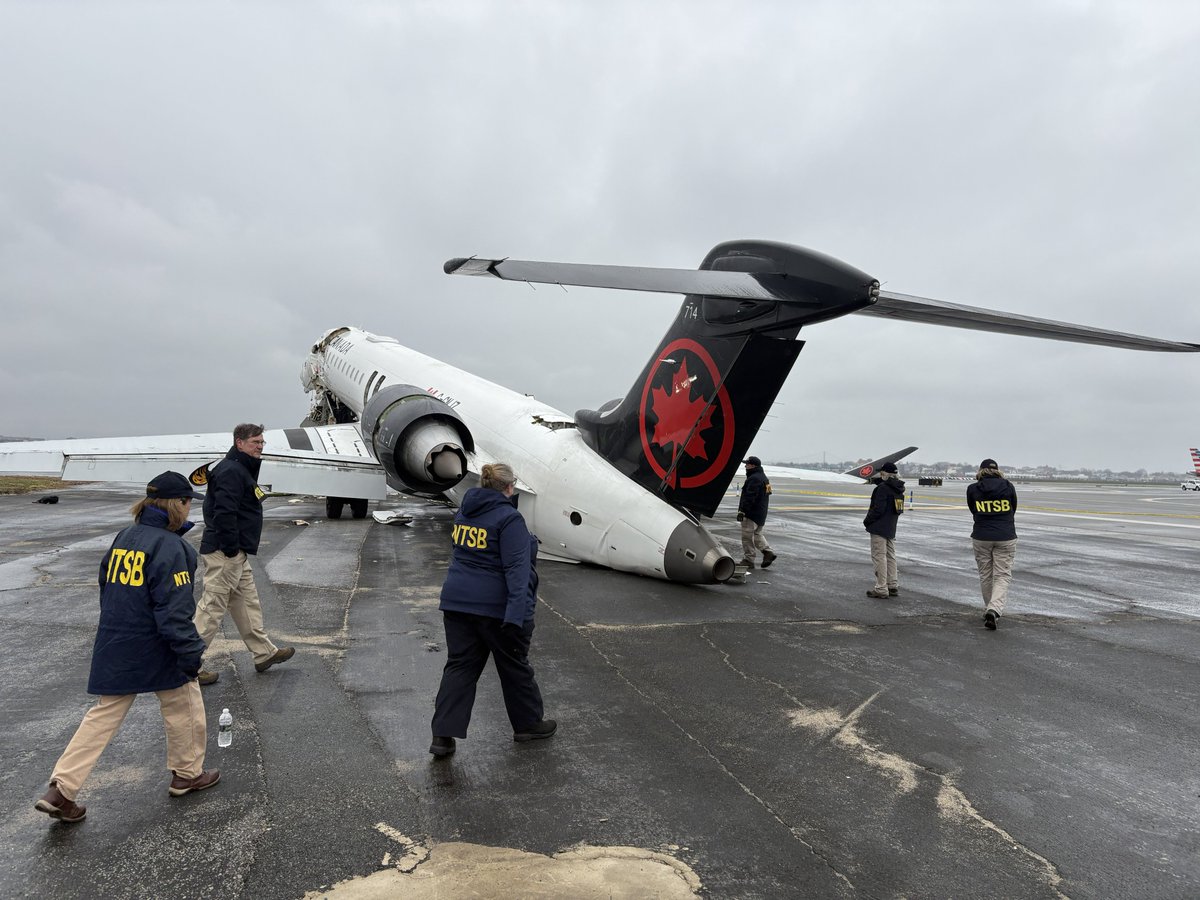 The image depicts a damaged airplane on a runway under a cloudy sky. The airplane is white with a prominent red maple leaf logo outlined in black on its tail, identifying it as an Air Canada aircraft. The tail of the airplane is tilted high off the ground while the nose appears to be damaged and resting on the runway. There are several people wearing jackets labeled "NTSB" in large yellow letters, indicating they are part of the National Transportation Safety Board. These individuals are inspecting the site, walking around the aircraft. The runway surface shows visible damage, with cracks and repaired patches. In the background, other aircraft and airport infrastructure are slightly visible, partially obscured by the overcast weather.