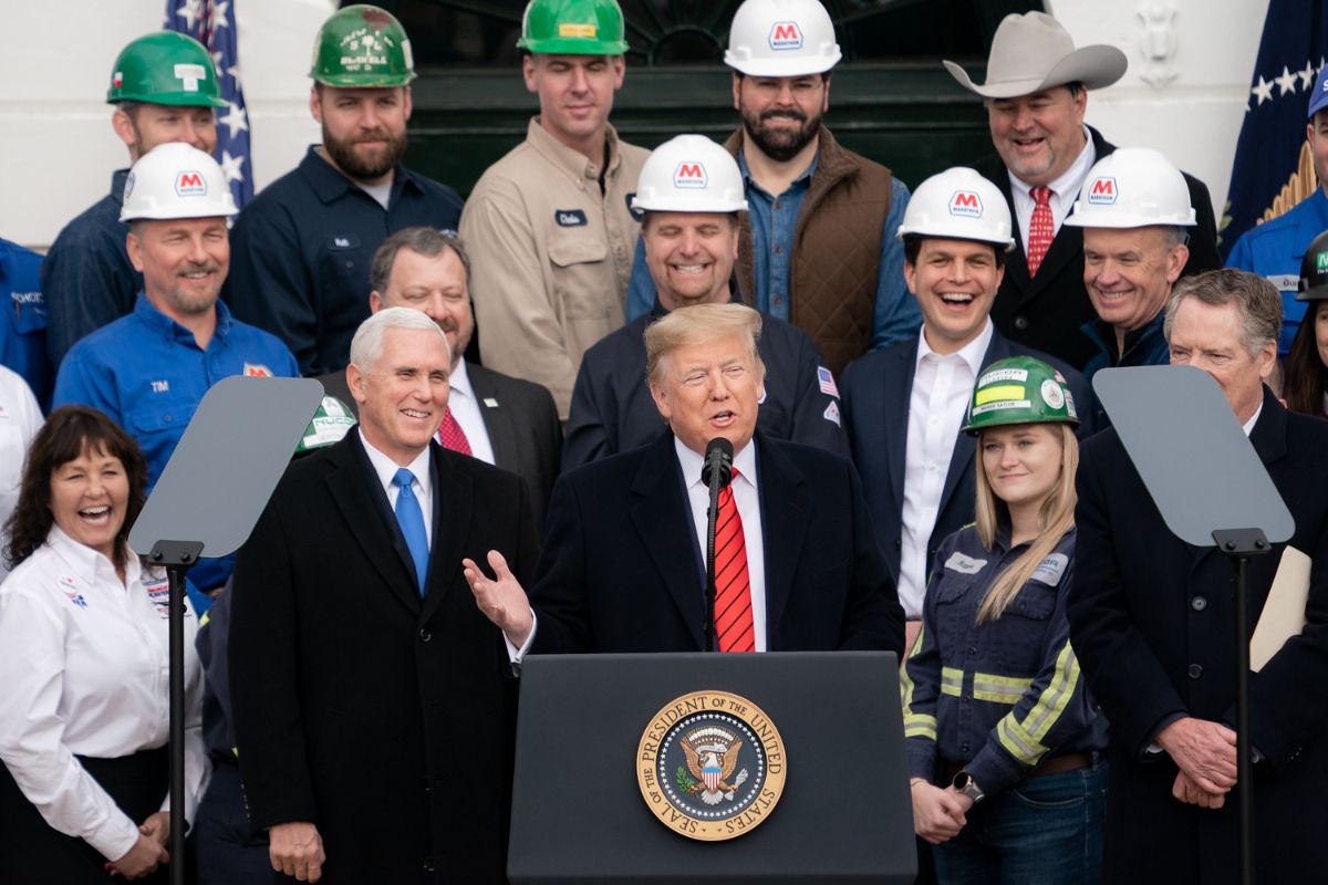 The image depicts a group of people standing outdoors, with a building in the background. There are multiple men wearing work uniforms, some with green and white hard hats labeled with various company logos. A central figure stands at a podium adorned with the seal of the President of the United States. The individuals appear to be smiling and engaged, with microphones and teleprompters visible. Flags are also partially visible in the background.