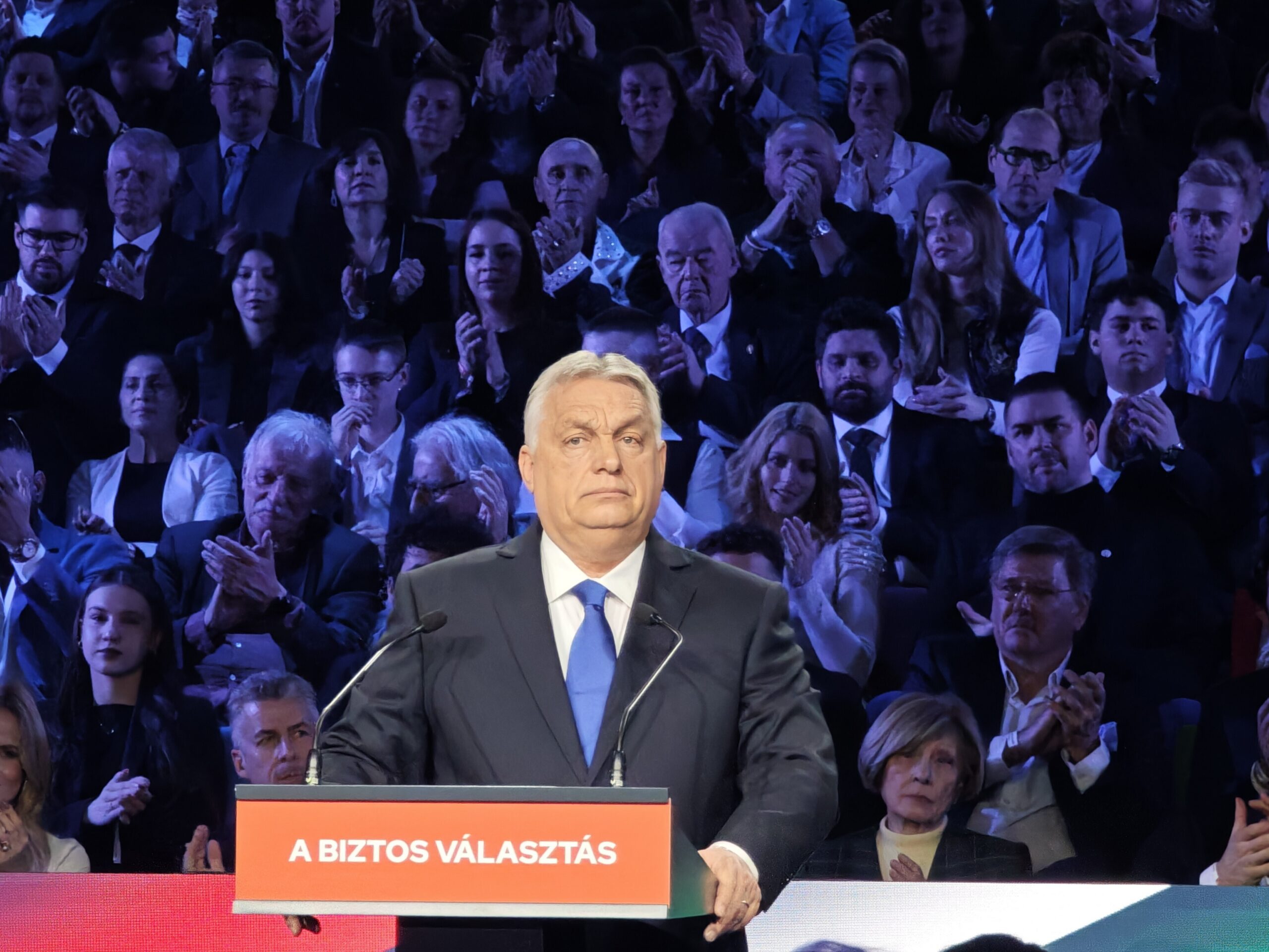 A public event where a middle-aged man in a dark suit and blue tie stands at a podium. The podium has a red sign with white text. He appears solemn and is positioned against a backdrop of an audience. The audience is densely packed and comprises various people in formal attire, some clapping and others observing intently. Low lighting casts a blue tint over the crowd, creating a dramatic atmosphere.