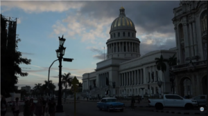 The image depicts a grand neoclassical building with a large domed roof, reminiscent of a capitol building. The structure features numerous white columns and a series of intricate designs along its facade. The dome is illuminated by soft light, contrasting against the darker, cloudy sky. In the foreground, there are several palm trees and an ornate street lamp with a curved arm. A classic blue car and a white vehicle are parked near the building. The scene has a twilight ambiance, with silhouettes of people walking near the street, providing a sense of scale and activity.