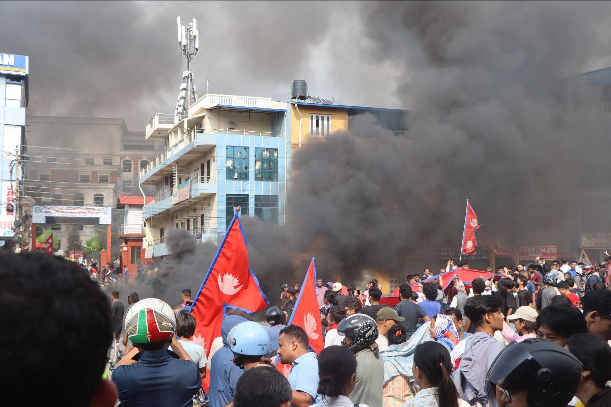 The image shows a crowded street scene with a large group of people gathered, some holding red flags with a white emblem. Thick black smoke billows heavily in the background, partially obscuring a modern multi-story building with blue glass panels and a visible rooftop telecommunications tower. The crowd consists of many individuals, some wearing helmets, indicating a potentially tense or hazardous situation. Amidst the crowd, various flags are held high, suggesting a protest or public demonstration. The dense smoke creates a stark contrast against the blue sky and the colorful scene below.