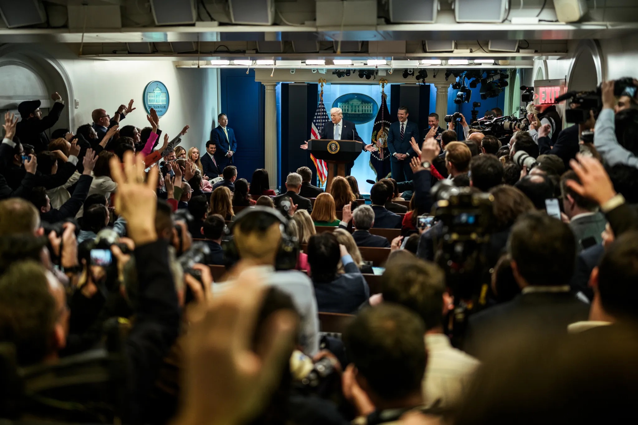 The image captures a busy press conference in a large room. At the front, a podium with the presidential seal is flanked by two flags—one with the United States flag and the other with the presidential flag. A speaker stands at the podium, with several people standing nearby, all in formal attire. The backdrop features a blue curtain and a logo of the White House. The room is filled with a large audience, predominantly journalists, many of whom have their hands raised. Numerous cameras are visible, pointing towards the podium. The crowd is seated, though several people are standing along each side. Overhead, lighting fixtures and panels are visible, providing illumination across the room.