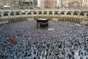 The image depicts a vast crowd of people gathered around the Kaaba, the cube-shaped building located at the center of the Grand Mosque in Mecca, Saudi Arabia. The Kaaba is covered with a black cloth adorned with gold-embroidered calligraphy. The crowd, mostly dressed in white, indicates a large number of pilgrims performing the Hajj. The surrounding architecture of the mosque features multiple levels with grand, arched walls and ornate detailing. In the background, modern skyscrapers can be seen rising above the mosque, juxtaposing traditional religious structures with contemporary urban development. The gathering is densely packed, illustrating the massive scale of the religious event.