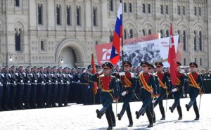 The image captures a military parade in front of a grand building with ornate architectural details featuring arches and decorative stonework. In the foreground, a group of uniformed soldiers marches in formation, each wearing dark green uniforms with red accents and gold epaulettes. Some soldiers carry flags, including a red flag and a flag with blue, white, and red stripes. The soldier leading the group is saluting. Behind them, a large formation of soldiers stands in a line, wearing blue uniforms. To the right, a large sign or banner displays partially visible text and imagery, hinting at a celebratory context.