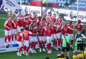 Soccer team celebrating with a trophy on a stage marked "CHAMPIONS 2025/26."