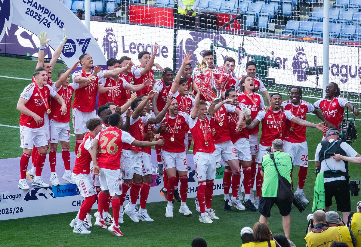 Soccer team celebrating with a trophy on a stage marked "CHAMPIONS 2025/26."