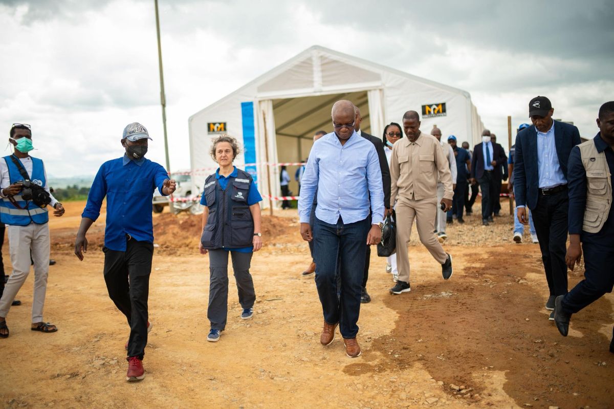 The image shows a group of individuals walking on a dirt pathway toward the camera. In the background, a large white tent structure with clear plastic walls is visible. The pathway appears to be part of a larger open space, possibly a field or construction site. The sky is overcast, with dense cloud cover. The individuals are dressed in a mix of formal and casual attire. Some are wearing blue vests and shirts, and various people are walking in different directions, suggesting an event or gathering.