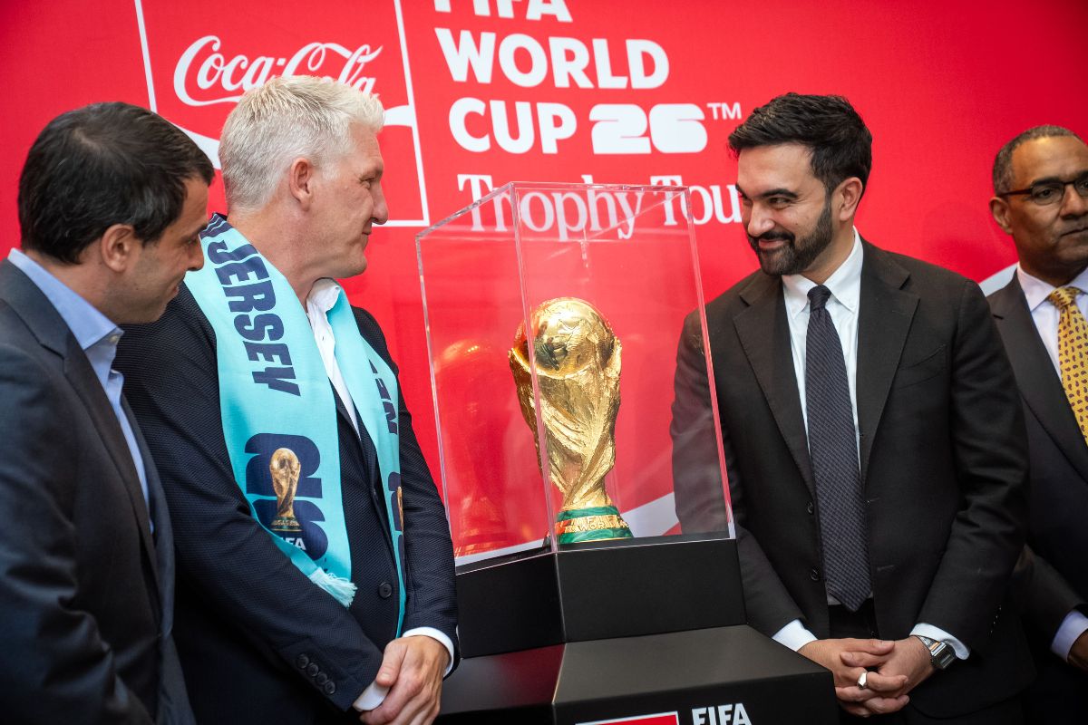 The image shows four men standing around a glass display case containing the FIFA World Cup trophy. The background features a vibrant red banner with white text. The man on the left is wearing a dark suit and light blue shirt. Next to him is a man with short white hair, wearing a blue suit with a blue jersey draped over his shoulder, which has "NEW JERSEY" printed on it. The third man is dressed in a black suit and tie with a light shirt. The man on the right wears glasses, a black suit, and a white shirt with a bright yellow patterned tie. They all look towards the trophy, which is displayed prominently in a clear case on a black pedestal.