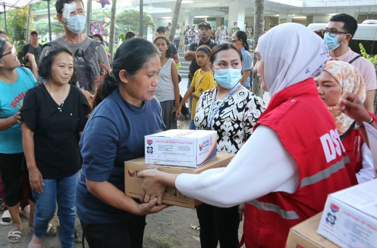 The image shows a group of people engaged in an aid distribution event. In the foreground, a woman in a blue shirt is receiving a cardboard box with printed labels from a person wearing a red vest, with "DSWD" partially visible on the back. The person handing over the box wears a white hijab, while another woman nearby is in a black and white patterned shirt with a face mask. More people are gathered around, some looking at the camera or the interaction taking place. In the background, there is a mixture of adults and children, with buildings and trees visible, suggesting an outdoor setting.