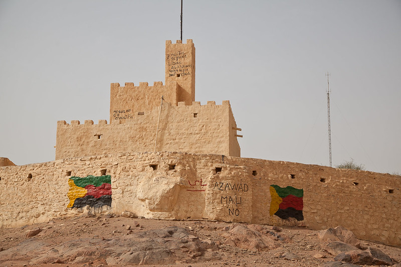 A sandy, textured fortress or building structure dominates the image. It is constructed from light-colored stone and features a series of rectangular battlements along the top. Below the main structure, there is graffiti featuring words and colorful symbols on the stone walls. The landscape around the building is barren, with rocky terrain and minimal vegetation. The sky is clear, adding to the arid atmosphere of the scene.