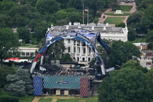The image depicts an aerial view of a large outdoor stage setup in front of a prominent white building with classical architecture. The building features columns and an ornate roof, partially obscured by a cluster of trees surrounding the area. The stage is adorned with an expansive metal arch structure decorated with red, white, and blue star patterns, suggesting a patriotic theme. The arch frames the front facade of the building and is fitted with lights and equipment for an event. Beneath the arch, a grid of assembled platforms and staging equipment is visible, with a few people milling about, appearing to prepare for or dismantle an event. To the side of the stage, there is a patterned backdrop composed of blue and red squares.