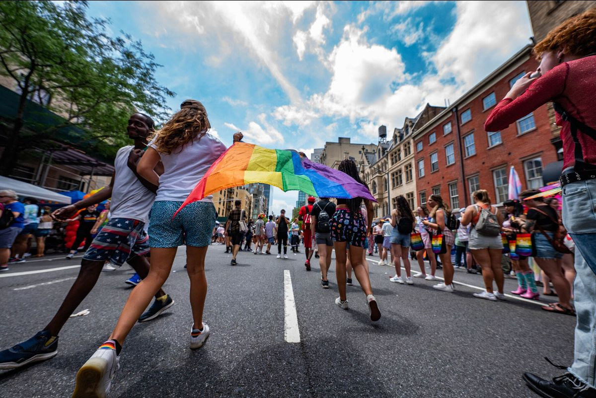 Two individuals run hand-in-hand, one holding a rainbow flag, among a crowd celebrating a Pride parade on a city street.