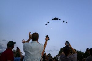 The image captures a sky scene during what appears to be an airshow or public event. In the foreground, a diverse crowd of people is seen looking upwards, some with their hands raised in applause and others holding mobile phones to record the spectacle. Their faces are illuminated by the fading light of dusk. Above them, a B-2 stealth bomber is prominently featured, flying in formation with several fighter jets, which are positioned below and to the side of the bomber. The sky is a gradient of soft blues transitioning into light gray, with a crescent moon visible. The overall composition reflects a moment of awe and excitement, characterized by the contrast between the aircraft and the natural scenery.