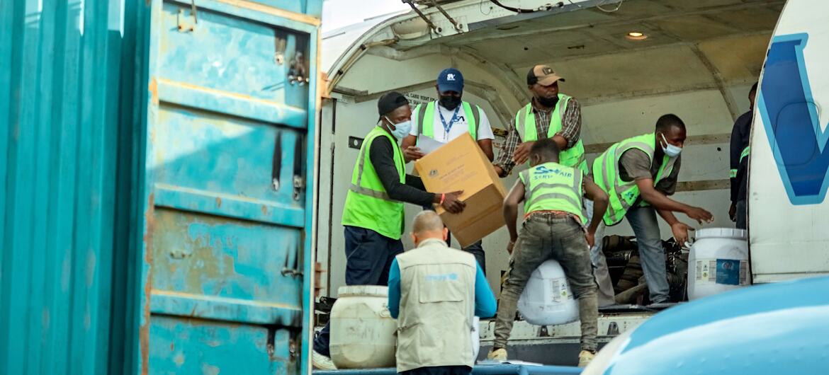 The image depicts a group of individuals engaged in unloading cargo from an aircraft. In the foreground, six people can be seen actively lifting and handling boxes and containers. They are wearing bright green safety vests and face masks, suggesting a focus on both safety and health protocols. The cargo area of the plane is visible, with various items, including large white containers and cardboard boxes, being unloaded. To the left, a section of a teal shipping container is partially visible, showing rust and wear on its surface, which adds context to the environment. The overall scene conveys a sense of urgency and collaboration in a logistics or relief effort setting.