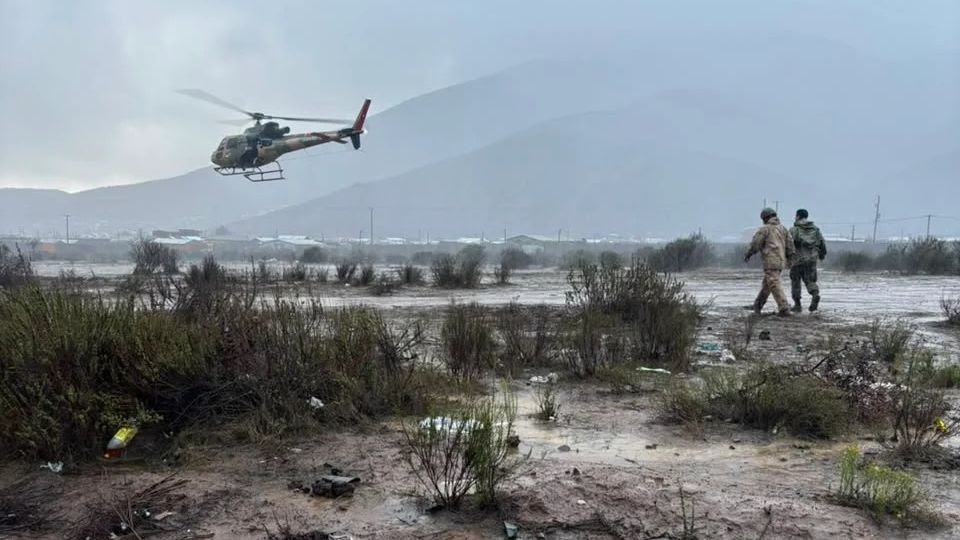 Military helicopter flying over two soldiers in a muddy landscape with sparse vegetation and mountains in the background.