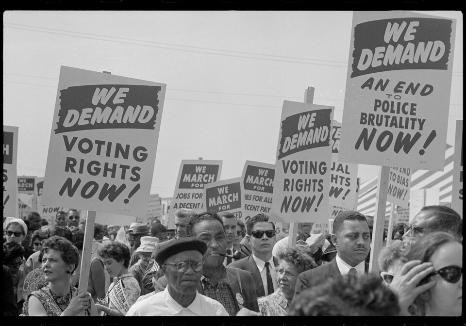 The image depicts a large crowd gathered in a public space, likely during a protest or demonstration. Many individuals are holding up signs with bold lettering. Prominent signs include statements such as "WE DEMAND VOTING RIGHTS NOW!" and "WE DEMAND AN END TO POLICE BRUTALITY NOW!" The crowd consists of a diverse group of people, including men and women of various ages. Some are dressed in formal attire, while others wear casual clothing. The scene is in black and white, emphasizing the seriousness of the occasion. The sky overhead is clear, contributing to a bright atmosphere despite the weighty subject matter.