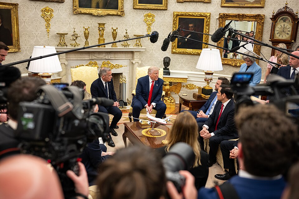The image features a meeting in an ornate room, likely the Oval Office. In the foreground, a group of individuals is seated around a central table, including a man in a dark suit and red tie, who appears to be the focal point of the discussion. The background displays gold-framed portraits and decorative items, adding to the room's luxurious feel. Several cameras and microphone booms are visible, indicating that this is a media-covered event. The individuals are engaged in conversation, with various expressions of attentiveness on their faces. Soft lighting from nearby lamps illuminates the scene.