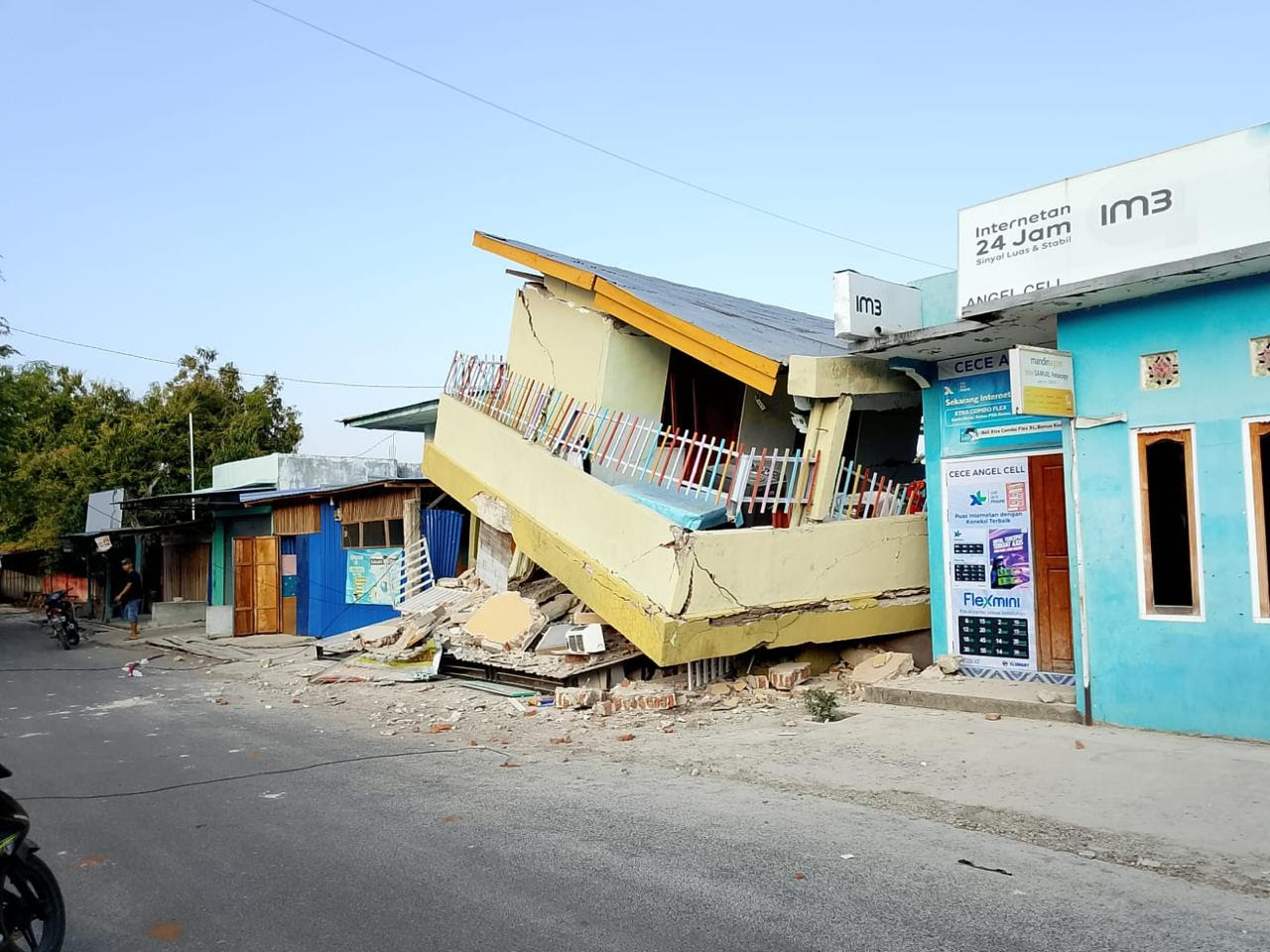 A leaning yellow building with structural damage from an apparent earthquake, alongside a blue building with signage, on an asphalt street.