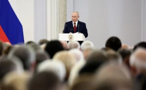 A man stands behind a white lectern, addressing an audience in a bright formal interior. He wears a dark suit, white shirt, and red tie, with two microphones positioned in front of him. A crest is displayed on the front of the lectern. A large Russian flag is partially visible along the left side of the image, while the foreground is filled with numerous out-of-focus audience members. White architectural columns and a pale brick-textured wall appear in the background.