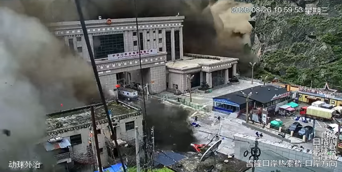 A surveillance-style image shows a gray, stone-faced multi-story building beside a steep, rocky, tree-covered mountainside. Large clouds of dark gray and tan smoke or dust obscure much of the left side and rise behind the building. The building has classical rectangular columns, broad terraces, decorative railings, and Chinese signage across its upper facade. In the foreground, a smaller structure appears damaged, with debris scattered across the ground and a leaning utility pole or cable crossing the scene. Several vehicles, including trucks and cars, are parked along a paved area on the right. Small storefronts or temporary structures with dark roofs, colorful awnings, and green umbrellas line the roadside. A few people are visible near the vehicles and buildings. White Chinese text overlays appear along the bottom and right edges, and a date-and-time stamp is displayed in the upper-right corner.
