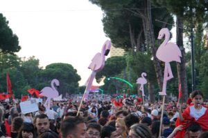 A large outdoor crowd fills a tree-lined avenue, with several pink flamingo-shaped signs mounted on wooden poles held above the people. The flamingos vary in size and position; some have black-and-pink wings, curved necks, and long legs. Red flags and banners are scattered throughout the crowd, and a child in a red-and-white outfit is being carried at the right edge of the image. Traffic lights, streetlights, and distant buildings are visible behind the crowd, while tall trees form a dense canopy along the roadway.