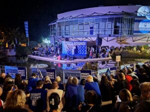 A nighttime outdoor political rally is shown in front of a modern, glass-fronted building illuminated with blue and purple lights. A raised platform spans a shallow reflecting pool, with speakers, microphones, musicians, and several people gathered onstage. A large blue backdrop displays Hebrew lettering and repeated logo-like text. A sizable crowd fills the foreground, many holding matching blue-and-white signs and flags. Additional signs line the barriers around the pool and stage. A large illuminated sign with Hebrew text and a portrait is mounted on the building at upper right. Trees, landscaping, and event lighting surround the scene.