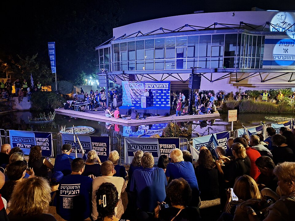 A nighttime outdoor political rally is shown in front of a modern, glass-fronted building illuminated with blue and purple lights. A raised platform spans a shallow reflecting pool, with speakers, microphones, musicians, and several people gathered onstage. A large blue backdrop displays Hebrew lettering and repeated logo-like text. A sizable crowd fills the foreground, many holding matching blue-and-white signs and flags. Additional signs line the barriers around the pool and stage. A large illuminated sign with Hebrew text and a portrait is mounted on the building at upper right. Trees, landscaping, and event lighting surround the scene.