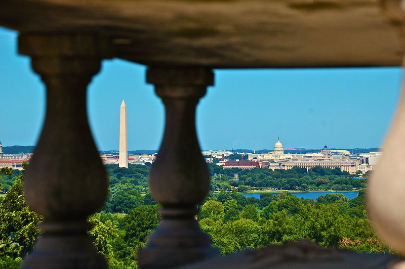 A cityscape is viewed through dark, weathered stone balusters in the foreground. Beyond the railing, dense green trees surround a narrow blue body of water, with the Washington Monument rising near the center-left and the domed U.S. Capitol visible in the distance to the right. Low civic and residential buildings extend across the horizon beneath a clear blue sky.