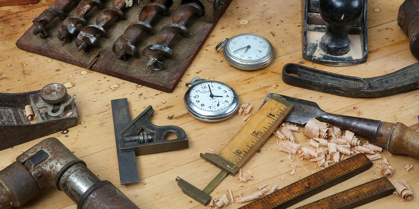 A close-up view of a worn wooden workbench covered with metalworking tools and hardware. In the upper left is a dark rectangular tray holding several rows of metal bits or punches. Two round dial gauges with black markings sit near the center, beside a black metal tool and a wooden measuring stick. A dark, rounded metal tool rests on a small rectangular plate near the top. Across the lower half are metal clamps, rods, fittings, a hammer-like tool, scattered wood shavings, and several elongated metal bars. The bench surface has scratches, stains, and visible wood grain.